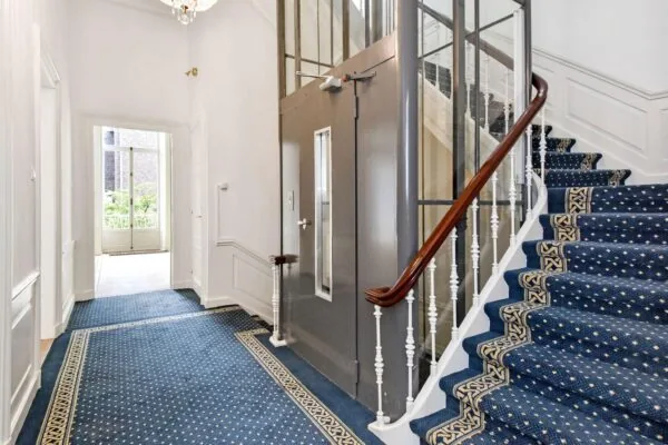 Statige hal in een herenhuis aan het Lange Voorhout met een klassieke trap, blauw tapijt en een moderne liftinstallatie.